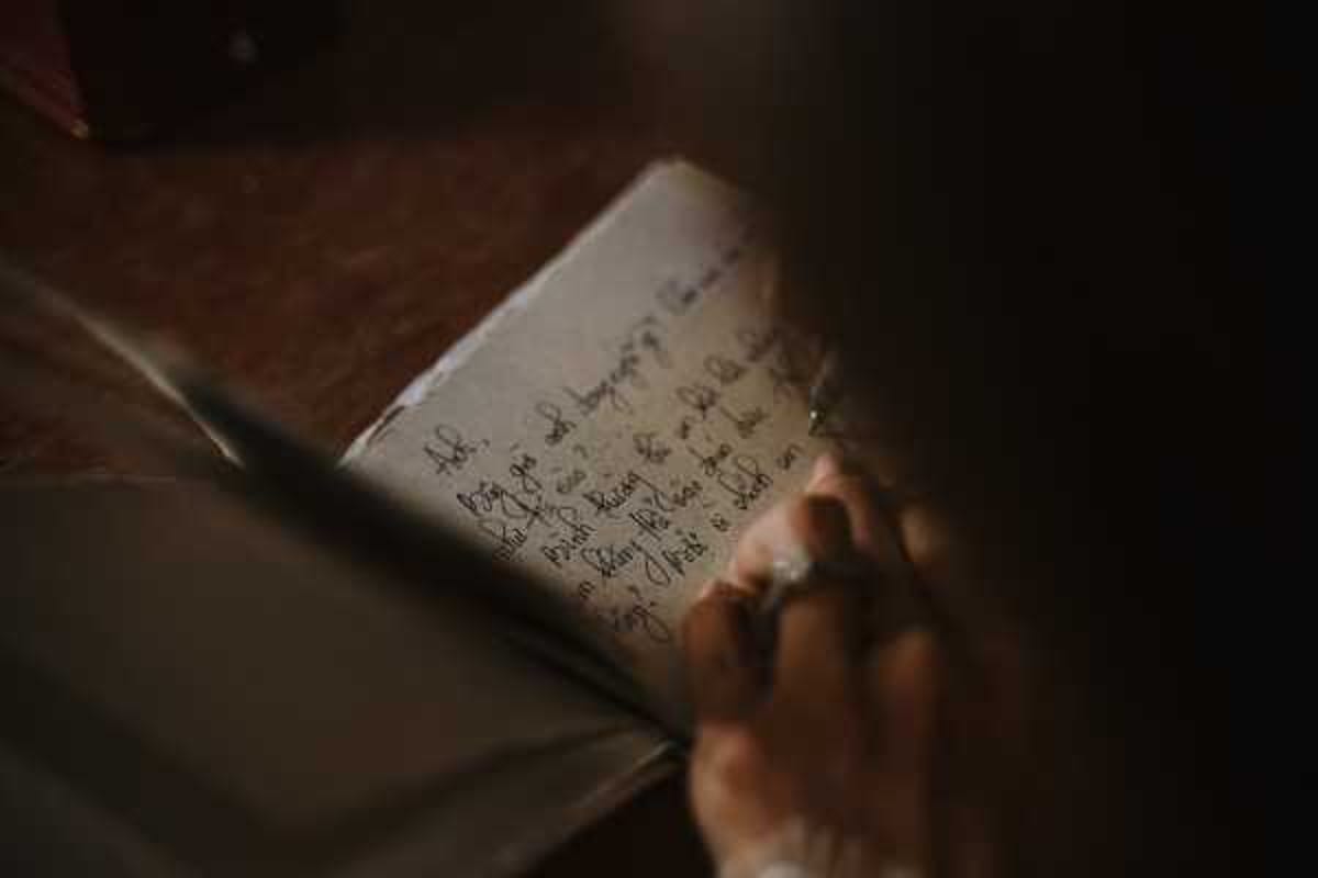A close-up detail shot of a person's hand holding a small notebook or journal open, with handwritten vows or notes visible on the pages in cursive script. The person appears to be wearing a ring, suggesting this may be a groom or bride reading personal vows before or during the ceremony. The image is shot in very low, moody ambient light with dark, shadowy tones and shallow depth of field, leaving much of the frame in soft blur. The overall palette is dark brown and warm shadow tones, with the written text serving as the focal point of the composition.