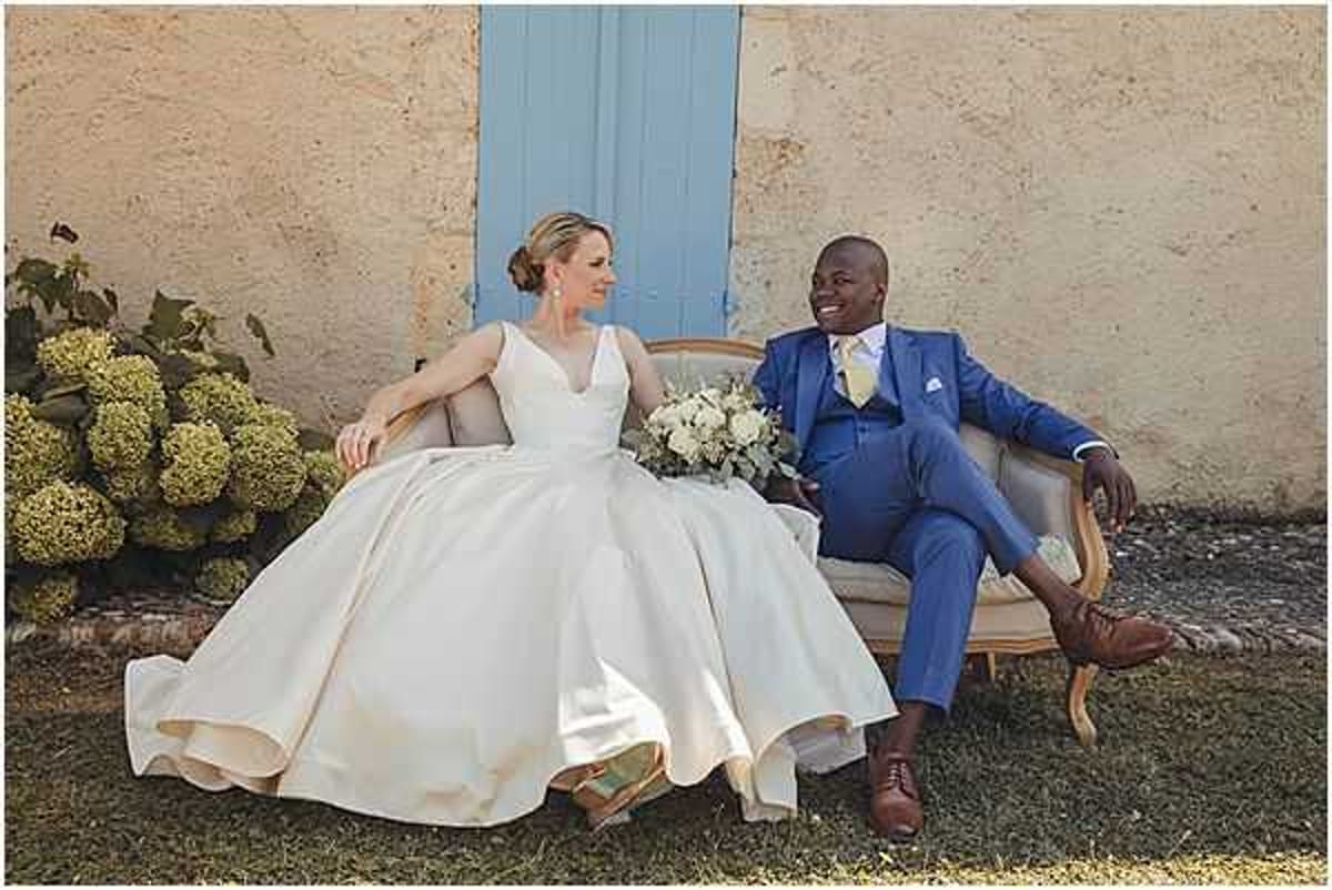 A couple portrait taken outdoors against a weathered pale ochre wall with a light blue painted shutter door. The bride wears a full-skirted, sleeveless ivory ball gown with a V-neckline and holds a bouquet of white roses and eucalyptus greenery. The groom wears a cobalt blue suit with a pale yellow tie and brown leather oxford shoes. The two are seated on a vintage-style upholstered settee with a gilded wood frame, facing each other and smiling. Lime-green hydrangea bushes are visible to the left. The overall styling has a classic French countryside feel. Medium-distance portrait shot with a slightly soft, natural light finish.