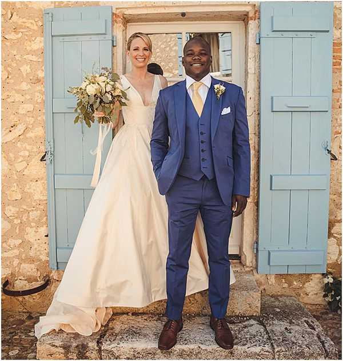 A couple poses together for a portrait outside a rustic stone building with pale blue wooden shutters. The bride wears an ivory ball gown with a deep V-neckline and holds a loosely arranged bouquet of cream roses, dusty blue thistles, and trailing eucalyptus with ivory ribbon streamers. The groom wears a cobalt blue three-piece suit with a gold tie, white pocket square, and a small floral buttonhole. Both are smiling directly at the camera. The shot is a medium full-length portrait framing both figures against the stone facade and blue shutters.