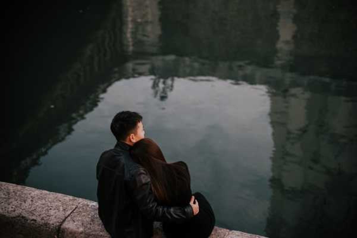 A couple sits together on a stone ledge beside a calm body of water, both dressed in black leather jackets, with the woman leaning her head against the man's shoulder as he holds her close. The shot is taken from behind at a slightly elevated angle, making the still, dark reflective water the dominant background element. The overall color palette is very dark and moody, with deep teals and near-blacks throughout. This appears to be an engagement or couples portrait session rather than a wedding day image, with a medium-distance portrait composition.