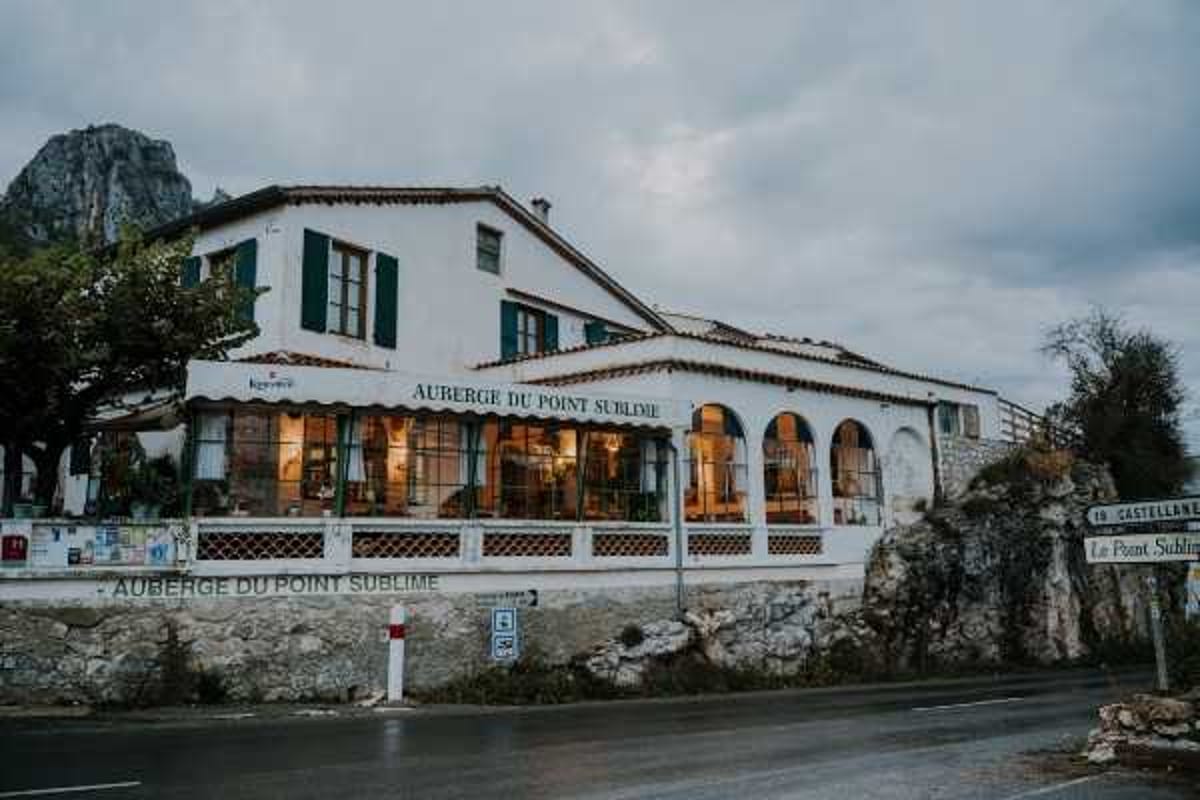 Wide exterior shot of the Auberge du Point Sublime, a white two-story French auberge with dark green shutters, arched windows along the ground floor, and a covered terrace with warm interior lighting visible through the glass. The building features traditional tiled roofing and whitewashed walls, with road signage for Castellane and Le Point Sublime visible in the foreground. No people or wedding party are present in the frame. Potential venue feature image.