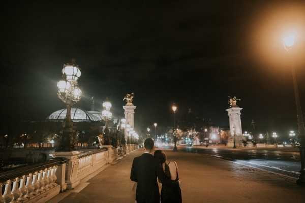A couple shares an intimate moment on the Pont Alexandre III bridge in Paris at night, photographed from behind in a wide shot. The groom is dressed in a dark suit and the bride wears a dark dress, their heads leaning together as they face the illuminated bridge stretching ahead of them. The ornate lamp posts and gilded sculptures of the bridge are lit up, with the glass dome of the Grand Palais visible in the background. The overall image has a dark, moody atmosphere with warm amber artificial lighting from the bridge lanterns and street lights.