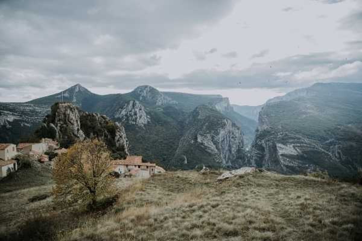 Wide landscape shot showing a dramatic mountain setting with a small cluster of traditional stone and terracotta-roofed village buildings perched on a hillside in the lower left of the frame. No people, couple, or wedding activity are visible in this image. The shot appears to be a location or venue establishing image capturing the rugged terrain and remote alpine village surroundings. Potential venue feature image.