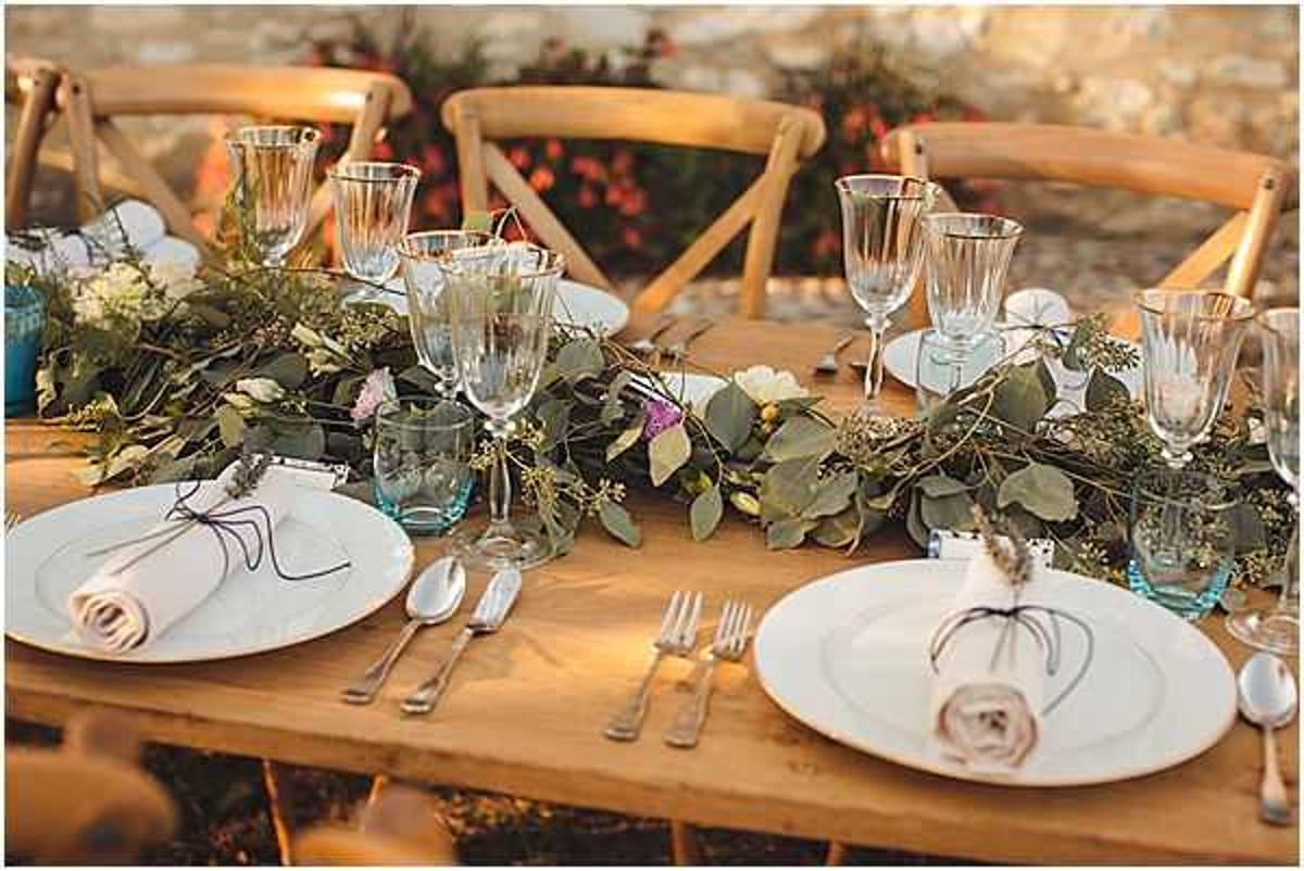 Close-up detail shot of an outdoor wedding reception tablescape set on a natural wood farm table with matching cross-back wooden chairs. A lush greenery garland runner made of eucalyptus, trailing vines, and small wildflowers in white and pale purple runs down the center of the table. Place settings include white round plates with linen napkins tied with twine and small dried flower accents, silver cutlery, and an assortment of clear and pale blue-tinted glassware including wine glasses and tumblers. The styling is rustic-bohemian with an earthy, natural palette.