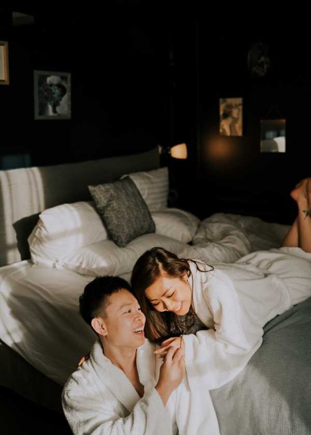 A couple relaxes together in a darkly decorated hotel or bridal suite bedroom, both wearing white robes, laughing and holding hands in a candid, intimate moment. The room features dark walls, a large bed with white linens and charcoal grey pillows, a warm-toned bedside lamp, and several framed artworks on the wall. The lighting is warm and moody, with a shaft of natural light illuminating the couple against the dark background. This appears to be a getting-ready or morning-after portrait, shot at close to mid range.