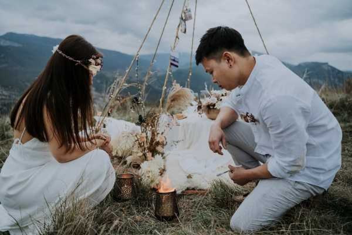 A couple is seated on the ground at an outdoor elopement or styled shoot set up on a mountain hilltop, both leaning in toward a small lit candle lantern. The bride wears a white off-shoulder dress and a delicate floral crown with dried burgundy and blush blooms, while the groom wears a white linen shirt and light grey trousers. Between them is a boho-style ground arrangement featuring a white blanket, dried pampas grass, white dried florals, and additional bronze or gold metal lanterns. A macramé or rope teepee frame with hanging decorations serves as a backdrop, giving the scene a boho-rustic aesthetic. The shot is a medium portrait taken at ground level, capturing both figures in profile.