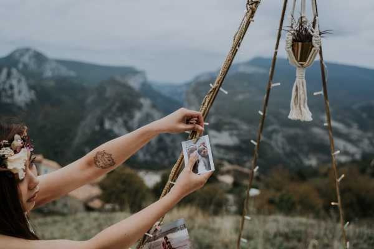 A bride attaches a Polaroid photograph to a rope-wrapped wooden teepee structure at an outdoor boho-style wedding, with a mountain landscape visible in the background. She wears a floral crown with dried and preserved blooms in muted pink and cream tones, and has a visible tattoo on her upper arm. The teepee structure is decorated with small peg-clipped photos and features a macramé hanging planter with dried botanicals in a brass pot. The shot is a close-up portrait taken from behind and to the side, emphasizing the decorative detail and the bride's interaction with the installation.