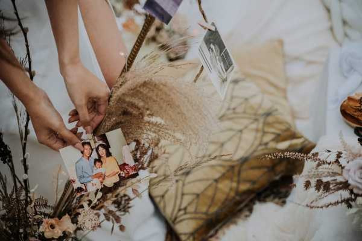 A close-up detail shot of a styled wedding flat lay arrangement on a white surface, featuring dried botanicals including pampas grass, dried ferns, and small blush blooms alongside a gold decorative pillow. Two hands are holding a family photograph showing what appears to be a family portrait with parents and children in casual clothing, suggesting a sentimental memorial or memory display element. Additional photographs and printed materials are visible in the background of the arrangement. The overall styling palette is warm and earthy with boho influences, incorporating dried neutral-toned florals as decorative elements.
