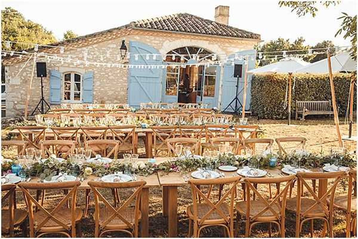 An outdoor wedding reception setup in the courtyard of a stone Provençal mas or farmhouse with pale blue shutters and terracotta roof tiles. Multiple long wooden farm tables are arranged in rows, lined with natural cross-back wooden chairs and dressed with lush greenery garland runners featuring white and soft-toned blooms, glassware, and white plate settings. String fairy lights and white triangle bunting are strung between posts above the dining area, and large white market umbrellas are visible to the right. The overall styling is relaxed rustic-Mediterranean with warm natural wood tones, greenery-forward florals, and a casual open-air feel. Wide establishing shot capturing the full reception layout. Potential venue feature image.