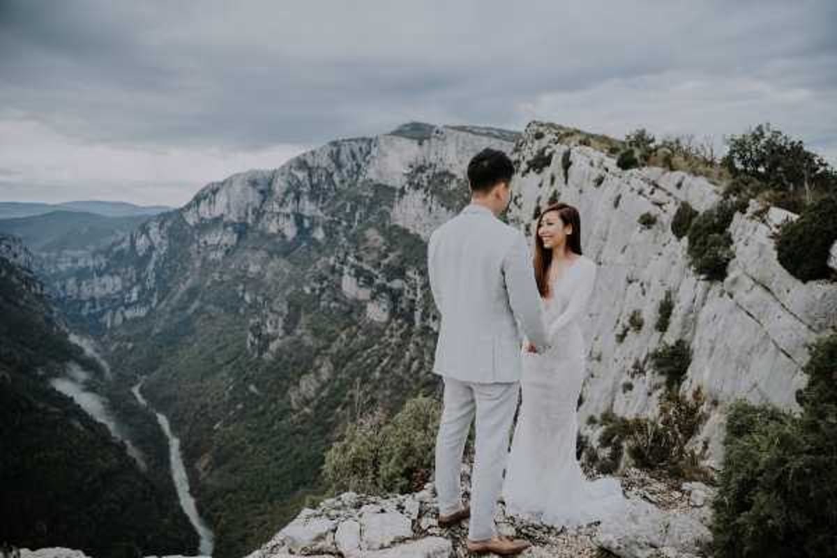 A couple poses together on a rocky cliff edge overlooking a dramatic canyon with a river winding far below, consistent with the Gorges du Verdon in southern France. The groom wears a light grey suit and the bride wears a long-sleeved fitted white gown; they are holding hands and facing each other. The shot is a medium wide portrait taken from behind and slightly to the side, with the vast canyon landscape serving as the backdrop under an overcast sky. The styling is minimal with no visible florals or additional decor, emphasizing an outdoor adventure or elopement aesthetic.