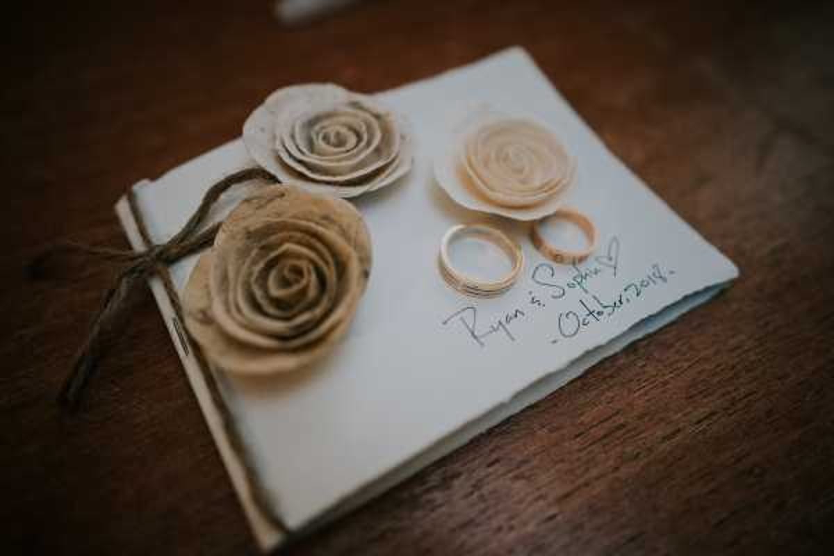 Close-up detail shot of a cream-colored wedding guest book or vow booklet placed on a dark wooden surface. The cover is decorated with three handmade rolled paper roses in ivory and aged-paper tones, tied with natural twine, giving a rustic aesthetic. Two gold wedding bands rest on the cover alongside handwritten text reading 'Ryan & Sophie - October 2018.' The composition is a shallow depth-of-field detail shot with warm, muted tones.