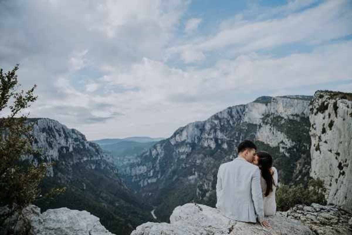 A couple sits together on the edge of a rocky cliff overlooking a dramatic canyon gorge, sharing a close, intimate moment with their faces nearly touching. The setting appears to be the Gorges du Verdon in Provence, France, shot as an outdoor portrait in a natural landscape. The groom wears a light grey suit and the bride wears a white or ivory dress, both facing away from the camera toward the expansive valley below. This is a wide-shot couple portrait with a boho or adventure-elopement aesthetic, emphasizing the scale of the natural surroundings against the two figures.