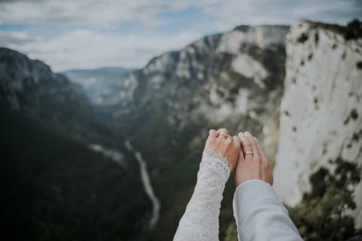 A close-up detail shot of a couple holding hands at an elevated outdoor overlook, with a dramatic gorge and river valley visible in the blurred background — consistent with the Gorges du Verdon in southeastern France. The bride's hand is adorned with a wedding ring and she is wearing a long-sleeved white lace dress, while the groom wears a light grey suit jacket. The composition is a tight portrait-style detail shot with the landscape dramatically out of focus in the background, emphasizing the joined hands as the focal point.