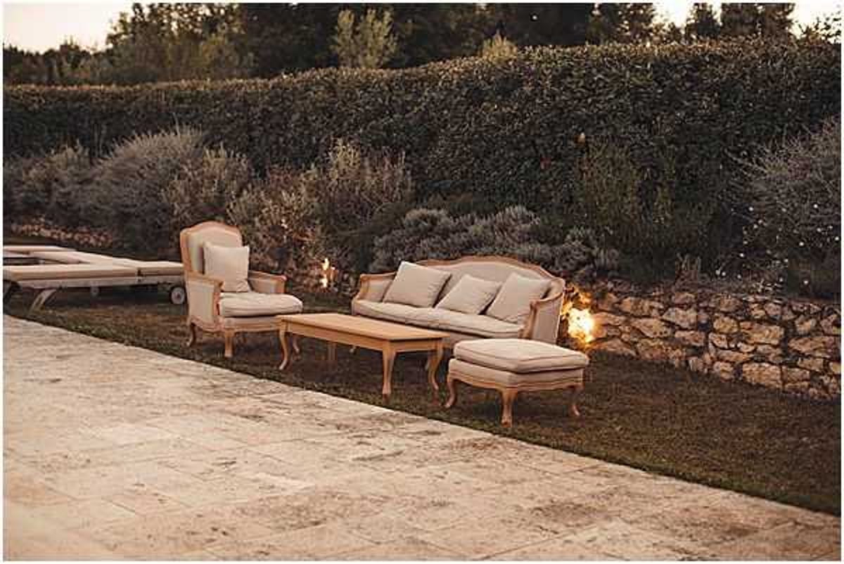 An outdoor cocktail hour or reception lounge area photographed at dusk, with no people visible. The seating arrangement consists of a French Louis-style sofa, a matching armchair, an ottoman, and a low wooden coffee table, all upholstered in neutral linen or beige fabric with carved wooden frames in a natural finish. Small candle lanterns placed on the ground provide warm ambient lighting along a low stone retaining wall. The setting is on a manicured lawn adjacent to a stone-paved terrace, with a dense hedge backdrop and lavender plantings contributing to a Provençal garden atmosphere. The overall decor style is classic French country. Wide establishing shot. Potential venue feature image.