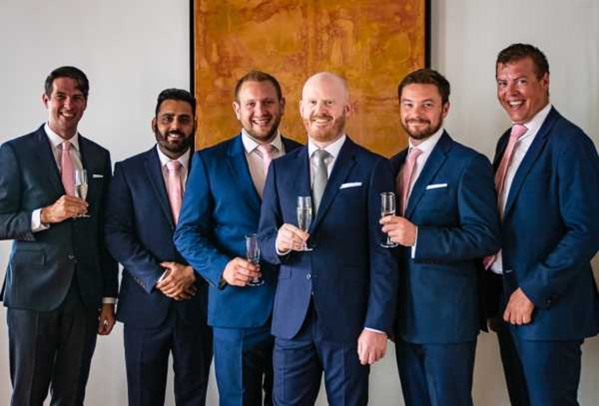 Six men pose together indoors in a getting-ready or pre-ceremony portrait, several holding champagne flutes. All six wear navy blue suits with light pink ties and white pocket squares, creating a coordinated groomsmen look. They are standing in front of a white wall with a large abstract painting in warm amber and orange tones. The shot is a medium group portrait with the men smiling directly at the camera.