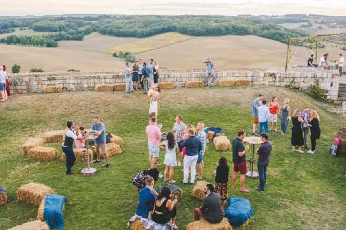 An outdoor cocktail hour taking place on a hilltop terrace or lawn with panoramic views of rolling agricultural fields in the background. Approximately 30-40 guests are mingling in small groups across the space. The seating and lounge areas are styled with hay bales arranged in clusters, some topped with blue fabric cushions, alongside tall cocktail tables with metal stands — a relaxed rustic theme. Guests are dressed in casual summer attire including bright colors and shorts, suggesting an informal dress code. The shot is taken from an elevated angle, likely from a balcony or upper floor, giving a wide overhead perspective of the entire cocktail area.