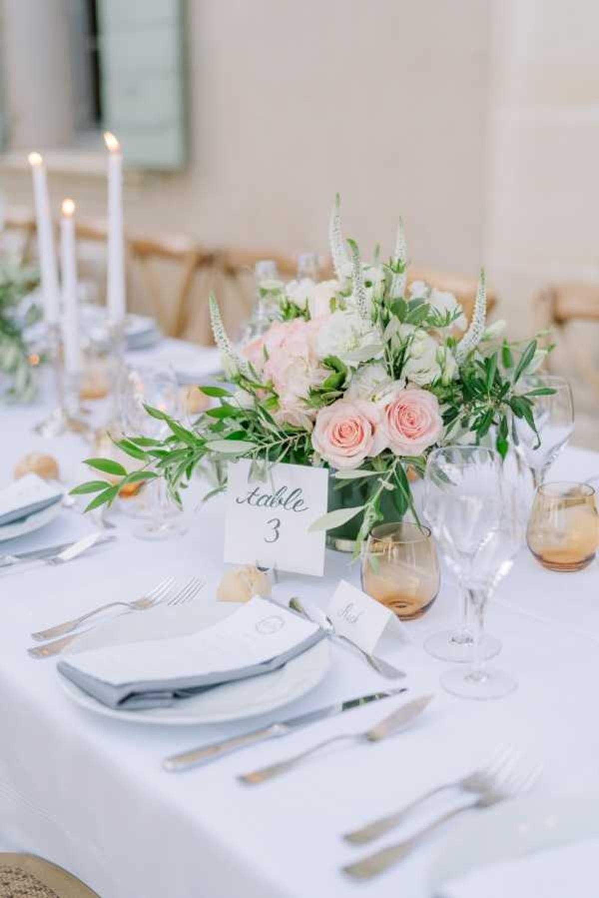 A close-up detail shot of a wedding reception table setting, likely at an outdoor or semi-outdoor venue. The table is dressed in a white linen tablecloth with place settings featuring white plates, grey folded napkins, silver cutlery, and amber-tinted glassware alongside clear crystal wine glasses. A low centerpiece of blush pink roses, white hydrangeas, white veronica spikes, and trailing green foliage sits in a clear glass vase at the center, accompanied by a calligraphy table number card reading 'Table 3' and small place cards. Tall white taper candles in candleholders are lit and visible in the background, contributing to a classic, soft color palette of blush, white, and sage green.