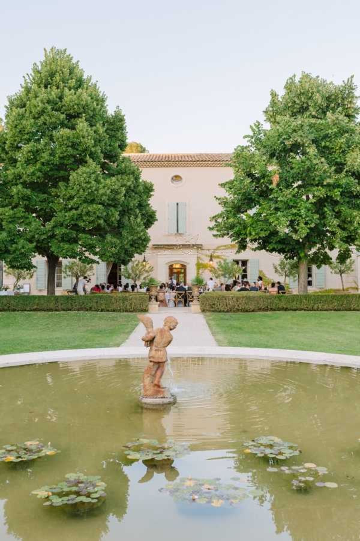 A wide outdoor shot of a wedding cocktail hour or reception taking place on the terrace of a large Provençal-style manor house with pale ochre rendered walls, blue-grey shutters, and a terracotta tile roof. A central gravel pathway leads from a circular ornamental pond — featuring a terracotta cherub fountain statue and floating lily pads — up to the gathering of approximately 30–40 guests seated and standing on the terrace. The setting is formal French garden style, with trimmed box hedging, symmetrically placed mature trees, and manicured lawn flanking the central axis. Potential venue feature image.