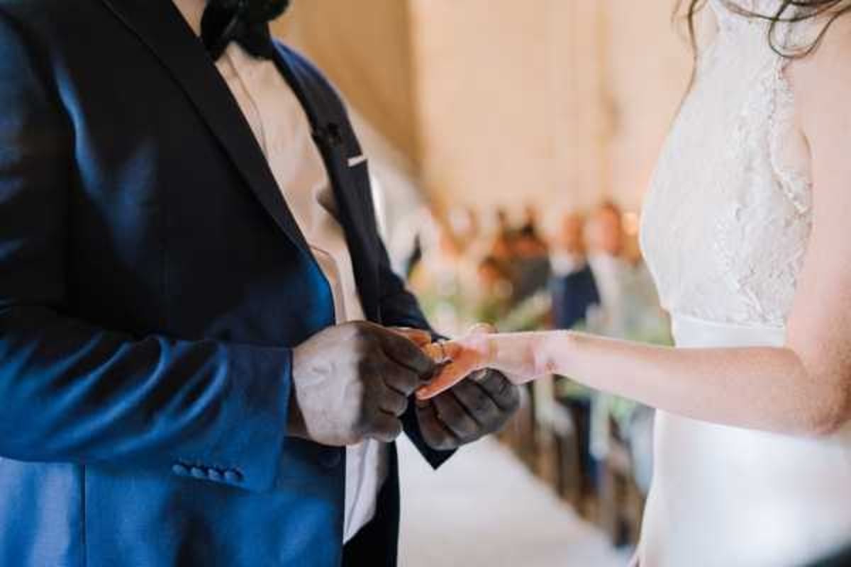 Close-up portrait shot of a ring exchange moment during an indoor wedding ceremony. The groom, wearing a navy blue suit jacket with a black bow tie and white shirt, is placing a gold band onto the bride's finger; the bride is wearing a white lace-detailed dress. Seated guests are visible in soft focus in the background, suggesting a chapel or stone-walled ceremony venue with warm natural light. The composition focuses tightly on the couple's hands, emphasizing the ring exchange as the central action.