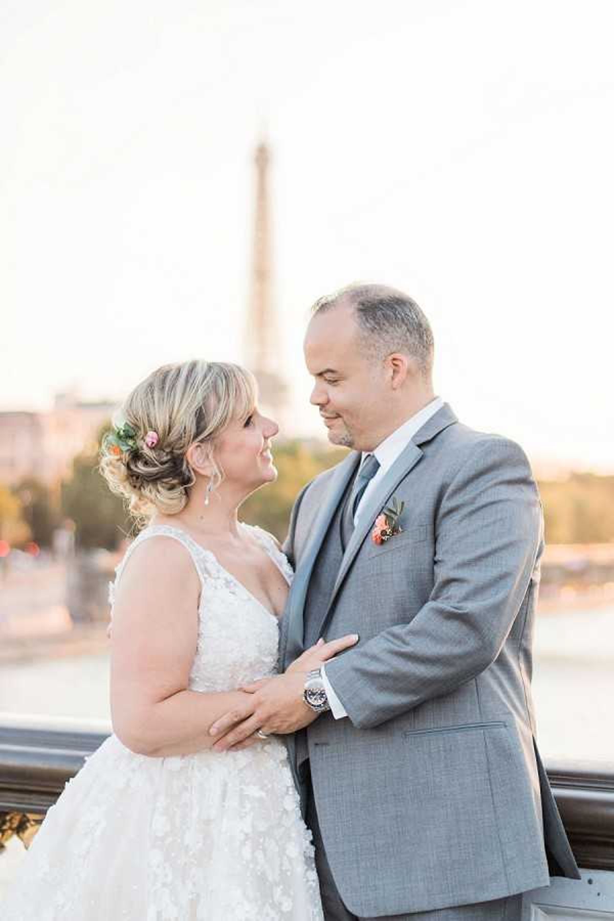 A couple portrait taken outdoors on a bridge in Paris, with the Eiffel Tower softly out of focus in the background along with the Seine River. The bride wears a sleeveless V-neck white lace ballgown with floral appliqué detailing, her blonde hair styled in a loose updo with small pink flowers tucked in. The groom wears a light gray three-piece suit with a gray tie and a boutonniere featuring coral and pink blooms with greenery. The two face each other closely with their arms around one another. The image is a mid-length portrait shot taken during golden hour light, giving the scene a warm, soft glow.