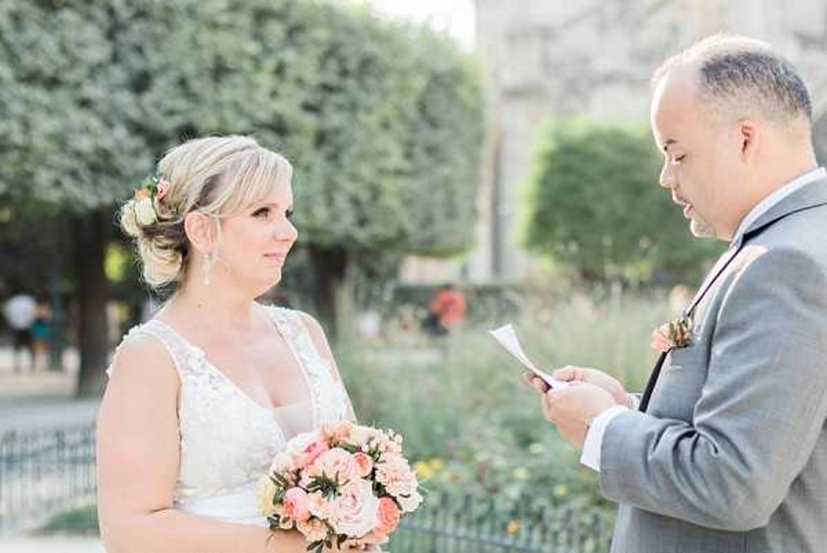 A couple shares an intimate vow exchange outdoors, likely in a French city park or garden setting. The groom, wearing a light grey suit with a floral boutonniere featuring peach and coral tones, reads from a small piece of paper while the bride looks at him attentively. The bride wears a lace V-neck white gown with her blonde hair styled in an updo adorned with small flowers, and carries a bouquet of peach, coral, and blush roses with soft greenery. The image is a medium close-up portrait shot with a shallow depth of field, with trimmed trees and an architectural structure softly blurred in the background.