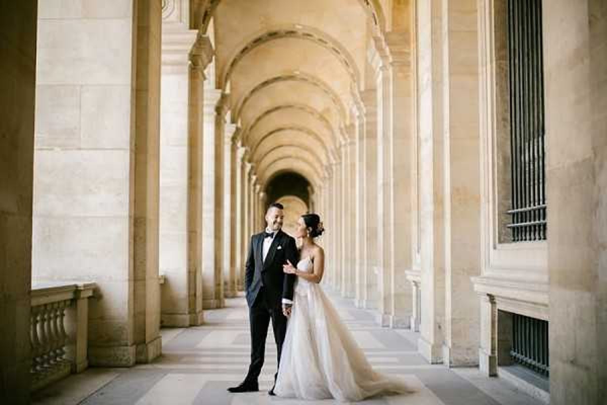 A couple poses for wedding portraits beneath a long vaulted arcade colonnade featuring repeating arched columns in pale limestone, consistent with classical Parisian architecture such as the Louvre or Palais Royal. The groom wears a black tuxedo with a bow tie, while the bride wears a strapless ballgown with a fitted embellished bodice and a voluminous layered tulle skirt in ivory. The two face each other and smile, captured in a wide shot that uses the receding colonnade to create strong architectural perspective and depth. The overall styling is classic and formal. Potential venue feature image.
