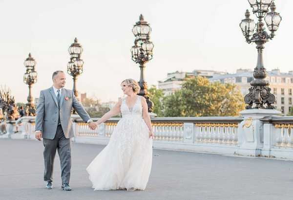A couple portrait taken outdoors on the Pont Alexandre III bridge in Paris, with the ornate bronze lamp posts and white stone balustrade clearly visible in the background along with Haussmann-style buildings. The bride wears a floor-length white ballgown with a deep V-neckline and lace appliqué detailing on the bodice and skirt, while the groom wears a light grey suit with a coral/blush boutonnière. The two are walking hand-in-hand across the bridge, the bride turned slightly toward the camera, shot in warm late-afternoon golden-hour light. The composition is a wide portrait shot with a soft, airy feel.