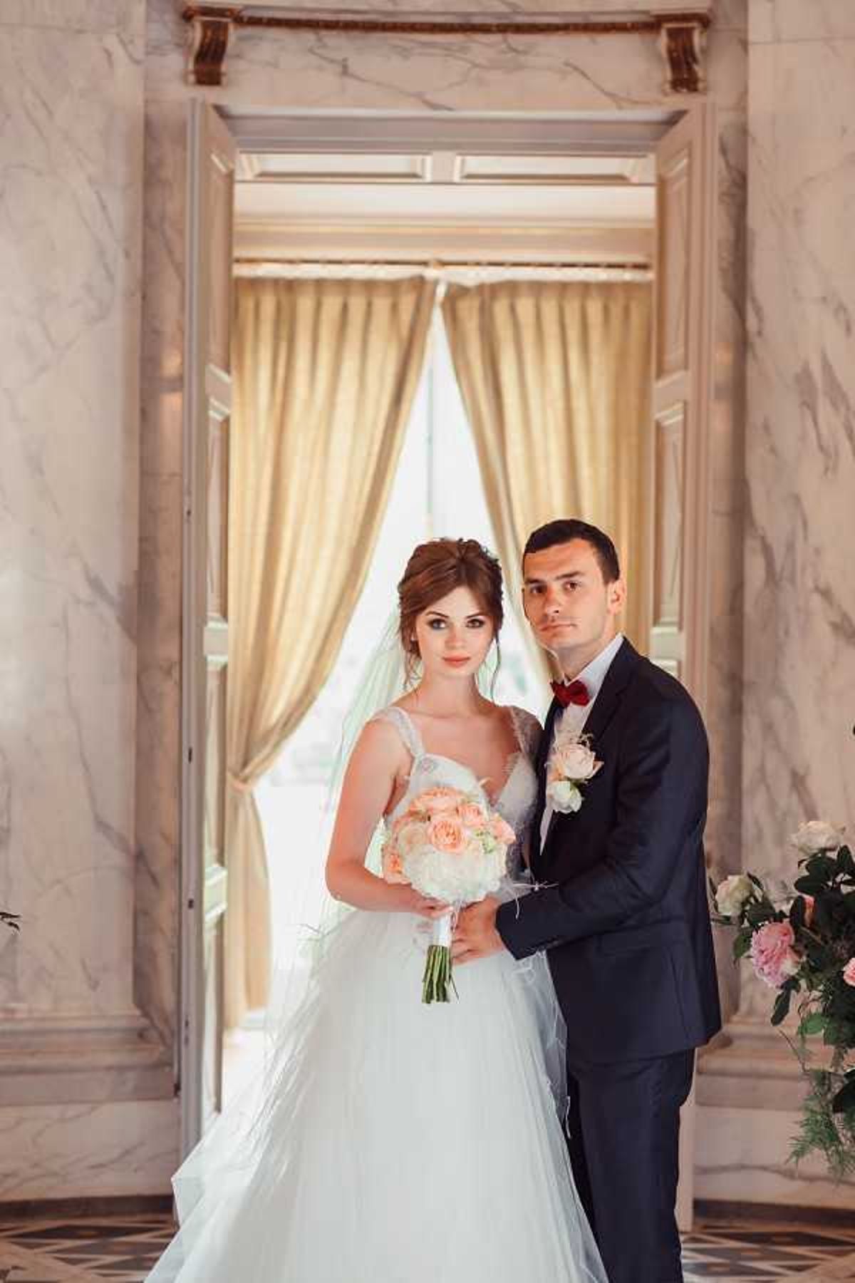 A couple portrait taken indoors in a formal venue featuring floor-to-ceiling marble walls and ornate doorframe architecture with gold floor-length curtains in the background. The bride wears a white ball gown with a lace bodice and a long veil, holding a rounded bouquet of peach garden roses and white peonies with greenery; the groom wears a navy suit with a burgundy bow tie and a white and pink rosebud boutonniere. Both face the camera directly, with the groom standing slightly behind and to the right of the bride, his arm around her waist. To the right edge of the frame, a partial arrangement of blush and pink roses is visible. The shot is a mid-length portrait with a classic, formal styling aesthetic. Potential venue feature image.