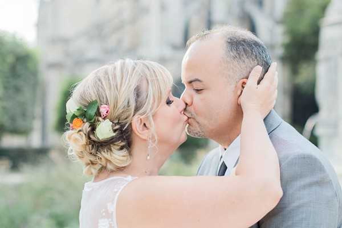 A close-up portrait of a bride and groom sharing a kiss outdoors, with a stone building visible and softly blurred in the background. The bride wears a white lace dress and has her blonde hair styled in an updo decorated with fresh flowers including pink roses, an orange bloom, and green foliage. The groom wears a light grey suit with a dark tie. The bride's hand is placed gently on the groom's face as they kiss. The image has a bright, airy feel with soft natural light.