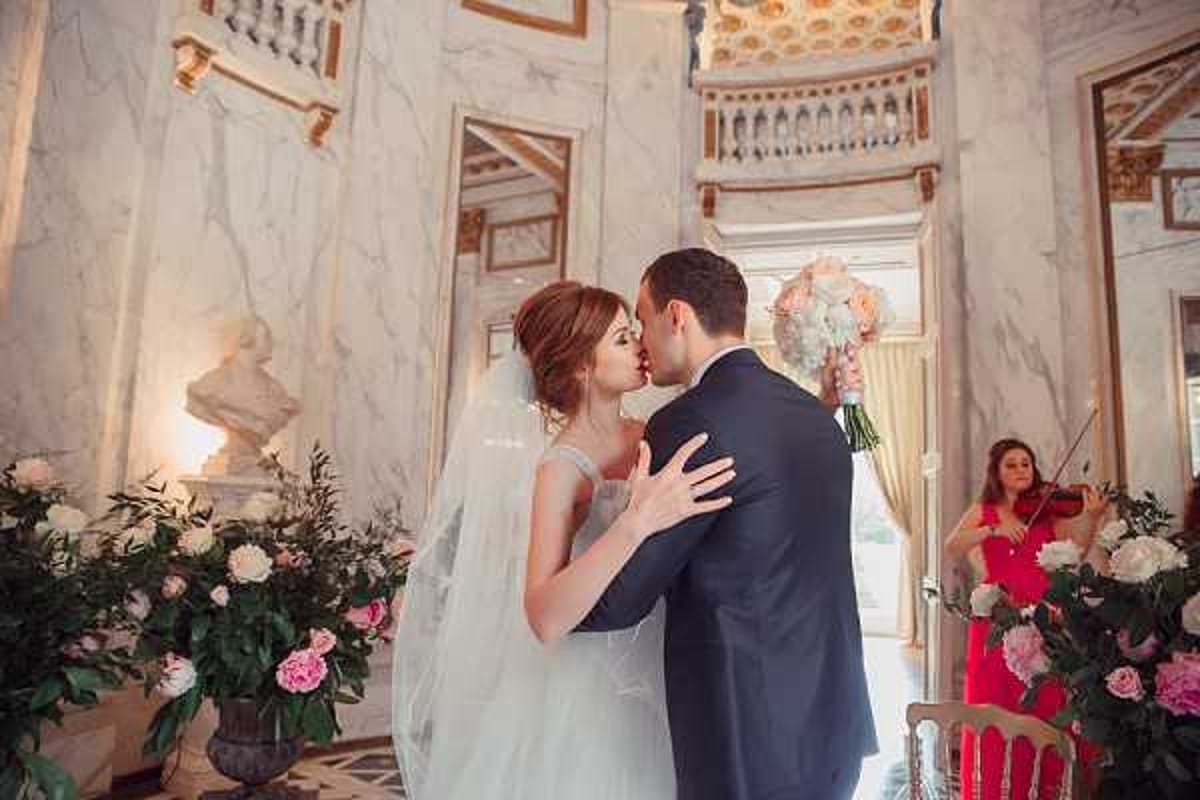 The bride and groom share their first kiss inside an ornate marble-clad hall of what appears to be a French chateau or palace, featuring gilded ceiling moldings, classical busts, and a grand balcony above. The bride wears a sleeveless ivory gown with a long veil, holding a bouquet of blush and white peonies with greenery, while the groom is dressed in a navy suit. Large floral arrangements of pink and white peonies in dark urns flank the couple on both sides, and a female musician in a red dress plays violin in the background to the right. The shot is a mid-length portrait capturing the intimate moment within the grand architectural setting. Potential venue feature image.