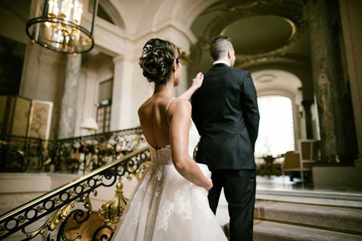 A couple portrait taken from behind as the bride and groom stand at the top of a grand interior staircase inside what appears to be a classical French palace or hôtel particulier. The bride wears a low-back, sleeveless ball gown with lace appliqué detailing and a beaded waistband, with her dark hair styled in an updo adorned with a decorative hairpiece; the groom wears a black suit. The staircase features ornate gilded wrought-iron balustrades, and the interior showcases arched ceilings, marble floors, and a gold chandelier overhead. The composition is a medium-wide shot from behind, emphasizing the architectural grandeur of the venue interior. Potential venue feature image.