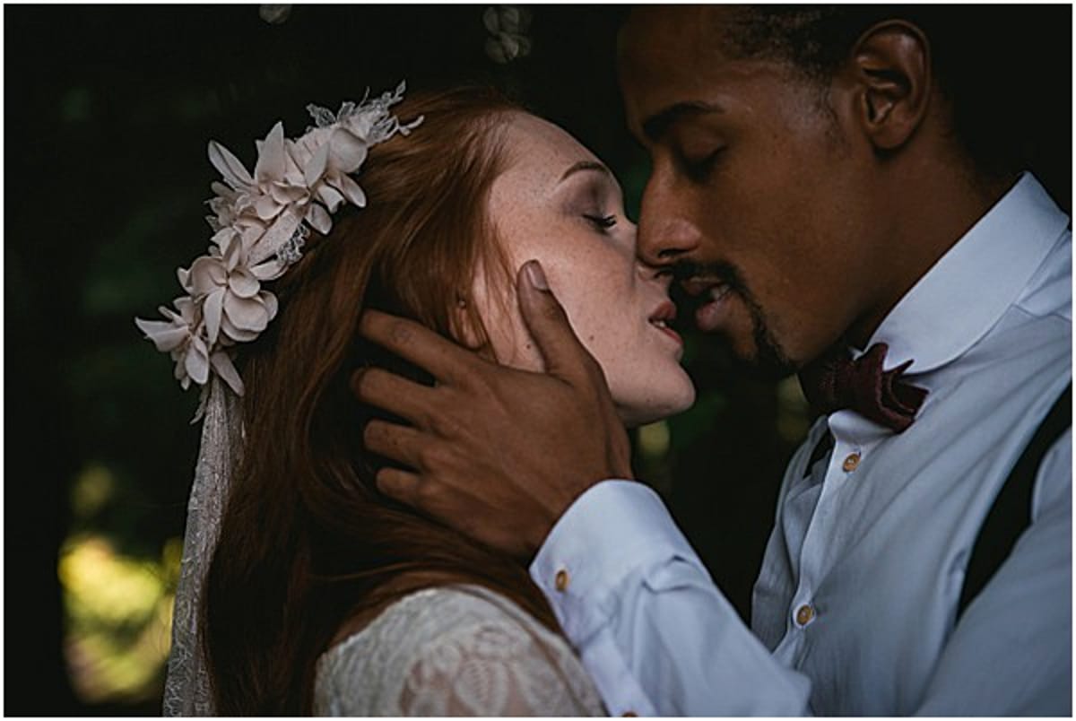 Close-up portrait of a bride and groom sharing a kiss outdoors against a dark, blurred green foliage background. The bride has long red hair and wears a floral headpiece made of cream fabric flowers with a veil, along with a lace dress. The groom wears a light grey suit jacket with a deep burgundy bow tie, and his hand gently cups the bride's face. The styling has a boho-romantic feel, with the floral headpiece and lace detailing as intentional design choices. The image has a moody, low-light quality with dark tones surrounding the couple.