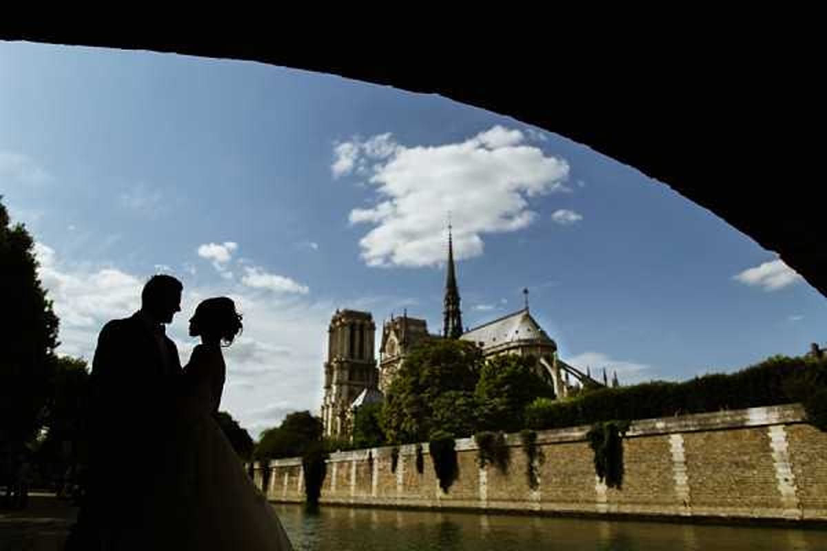 A couple is photographed as silhouettes beneath a bridge arch along the Seine River in Paris, with Notre-Dame Cathedral visible in the background. The composition frames the two figures — a bride in a full-skirted dress and a groom in a suit — facing each other in an intimate pose against a bright blue sky. The shot is taken from under the bridge, using the curved arch as a natural frame in the foreground, creating strong contrast between the dark silhouettes and the sunlit background. This is a wide environmental portrait emphasizing the Parisian landmark setting.