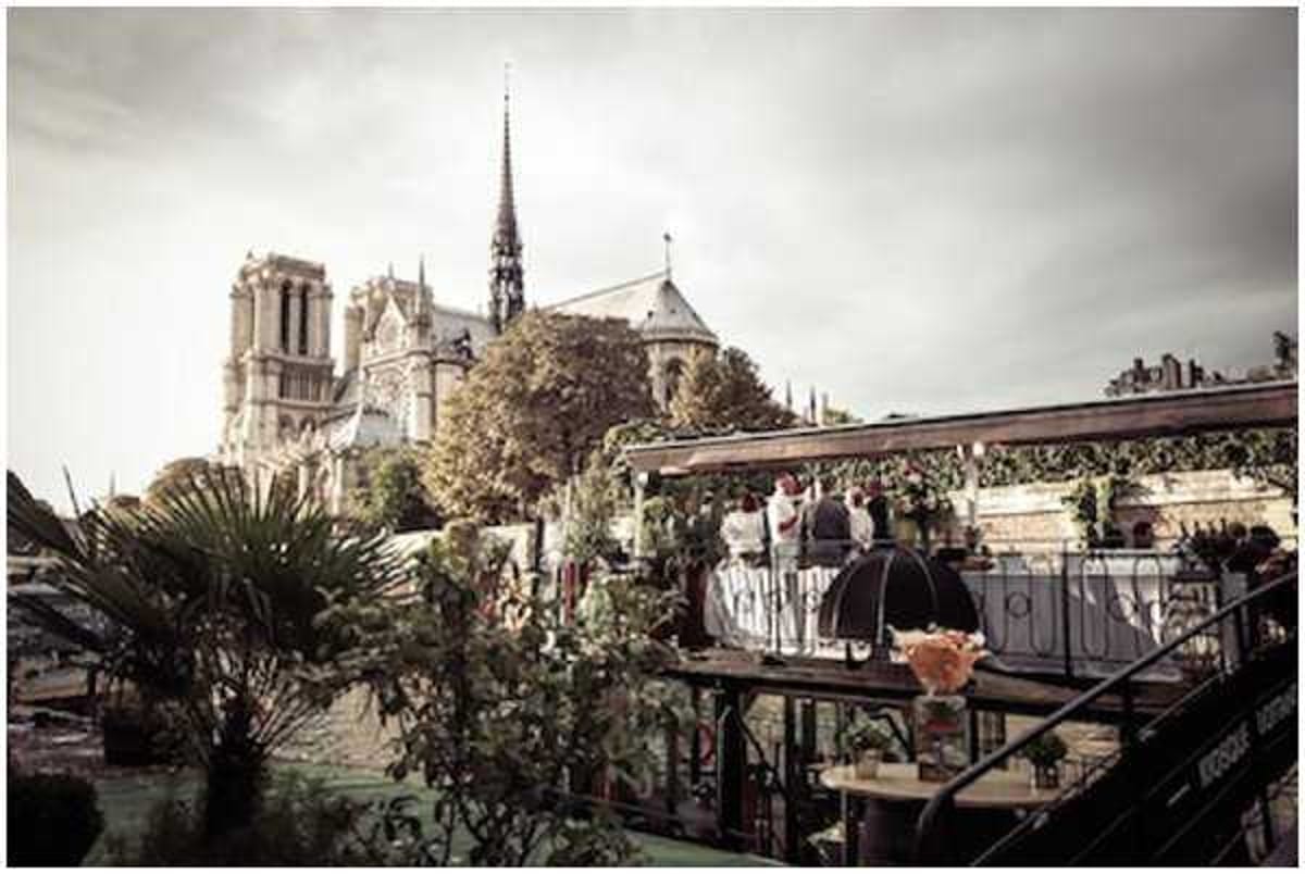 A wide-angle shot of what appears to be a wedding cocktail hour or reception taking place on the open-air deck of a barge or riverboat on the Seine in Paris, with Notre-Dame Cathedral prominently visible in the background. A small group of guests, approximately three to four people, are gathered along the decorated railing of the upper deck, which is adorned with lush greenery, potted plants, and trailing foliage creating a garden-like atmosphere. The deck features small round tables and dark iron railings, with the overall decor leaning toward a romantic garden style. The image has a moody, slightly desaturated tone due to overcast lighting conditions. Potential venue feature image.