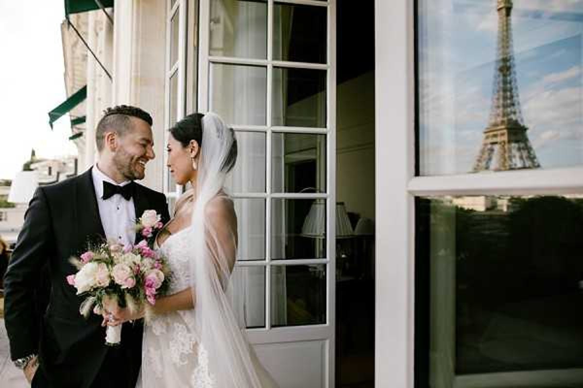 A couple poses together outdoors on a terrace or balcony in Paris, with the Eiffel Tower reflected in the window glass beside them. The groom wears a black tuxedo with a bow tie and boutonniere, while the bride wears a lace strapless wedding gown with a long cathedral-length veil. She holds a bouquet of blush pink and white blooms, including roses and what appear to be peonies, with light greenery. The two face each other closely and smile in a portrait-style mid shot, with the architectural facade of what appears to be a Haussmann-style building visible behind them.