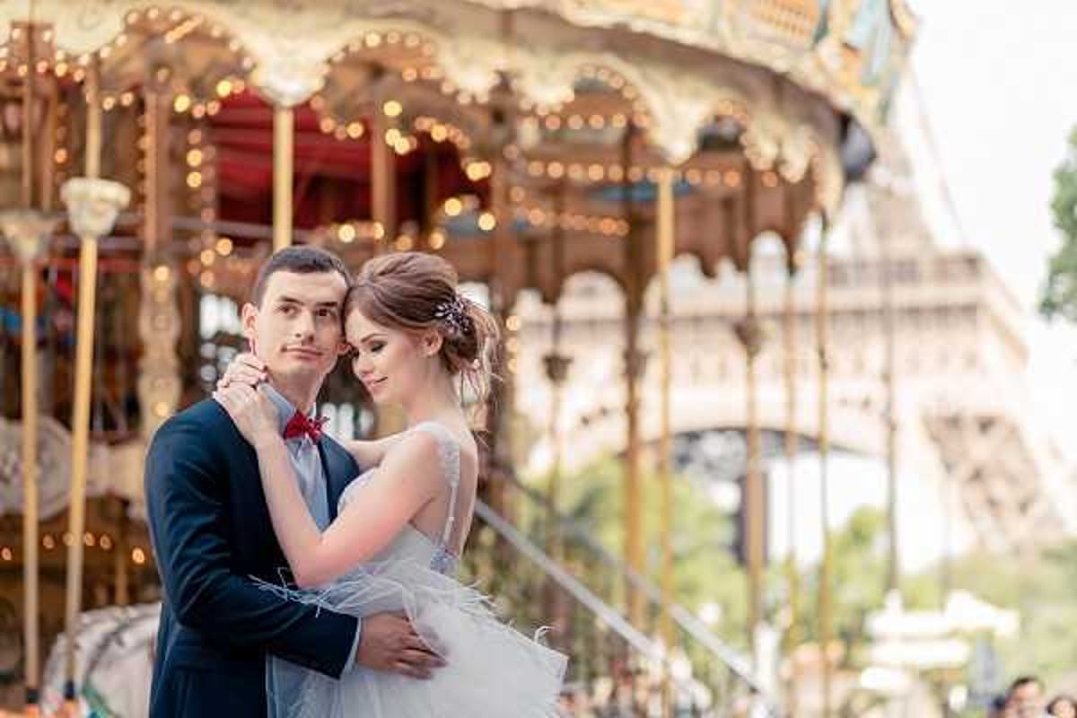 A couple poses for a portrait session outdoors in Paris, with the Eiffel Tower and a classic Parisian carousel visible in the background. The groom wears a navy suit with a red bow tie, while the bride wears a white gown with a feathered skirt hem and a sheer embellished bodice, her hair pinned up with a decorative hair accessory. The bride rests her arms around the groom's neck as he holds her close, both facing the camera in a relaxed embrace. The shot is a medium close-up portrait with the carousel's warm string lights and ornate gold detailing softly blurred behind them.