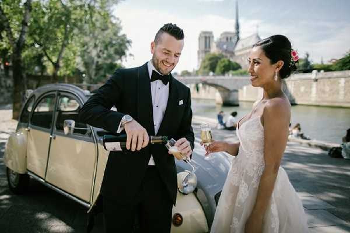 A couple portrait shot outdoors along the Seine River in Paris, with Notre-Dame Cathedral visible in the background. The groom, wearing a black tuxedo with a bow tie and white pocket square, is pouring champagne into a flute held by the bride, who wears a white lace spaghetti-strap gown and has a small pink flower in her dark upswept hair. They are standing beside a cream-colored vintage Citroën 2CV. The image is a mid-range portrait with natural daylight and a candid, celebratory mood.