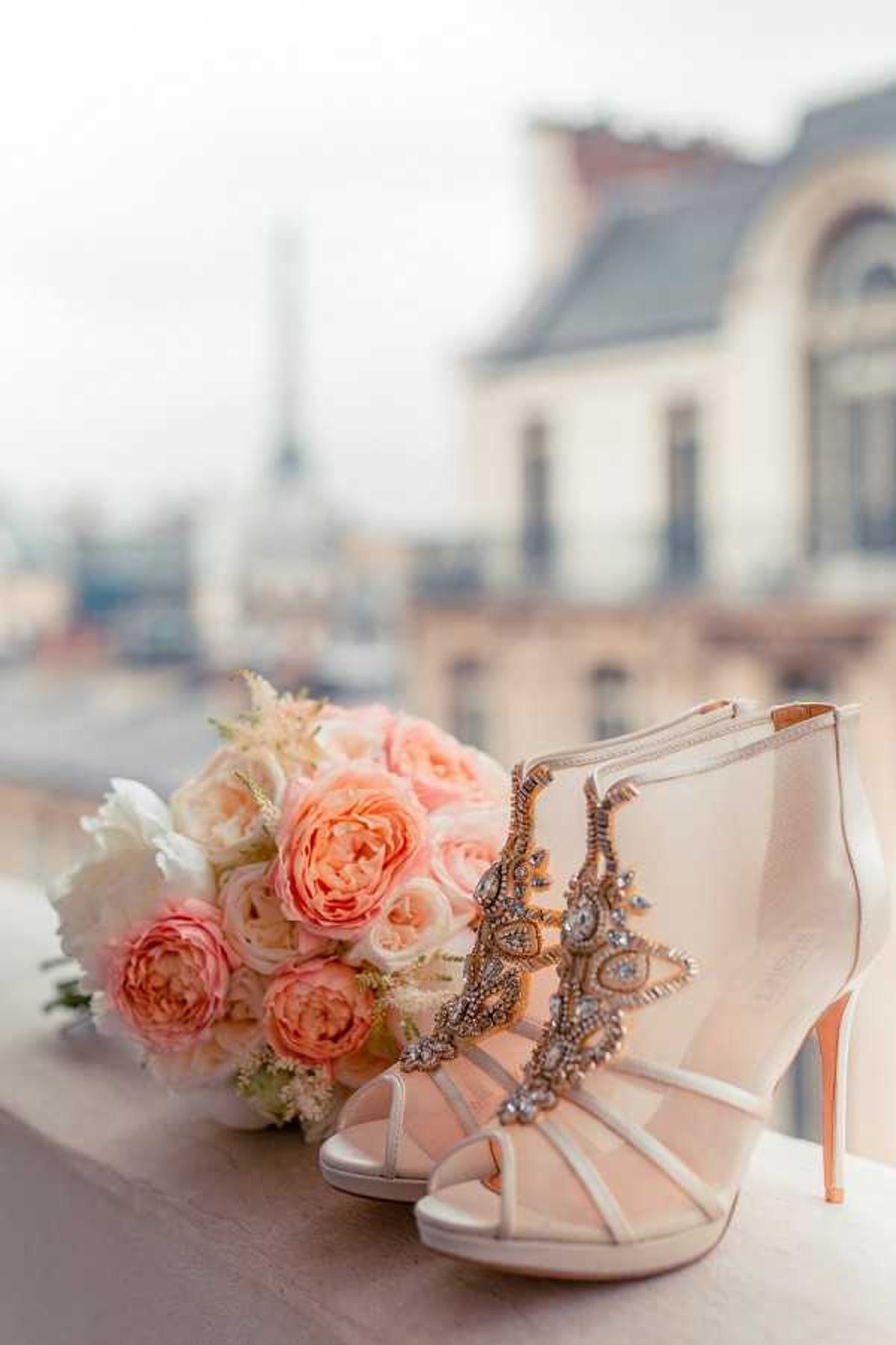 A close-up detail shot of bridal accessories arranged on a ledge or balcony railing with a blurred Parisian rooftop cityscape in the background, including what appears to be the Eiffel Tower in the far distance. The focal point is a pair of ivory strappy high-heel sandals with a rose-gold stiletto heel, featuring elaborate crystal and rhinestone embellishments along the vamp straps in an Art Deco-inspired pattern. Beside the shoes sits a bridal bouquet composed of coral and blush garden roses, white spray roses, and cream astilbe or similar filler flowers. The overall color palette is soft blush, ivory, and rose-gold, consistent with a classic Parisian wedding aesthetic.