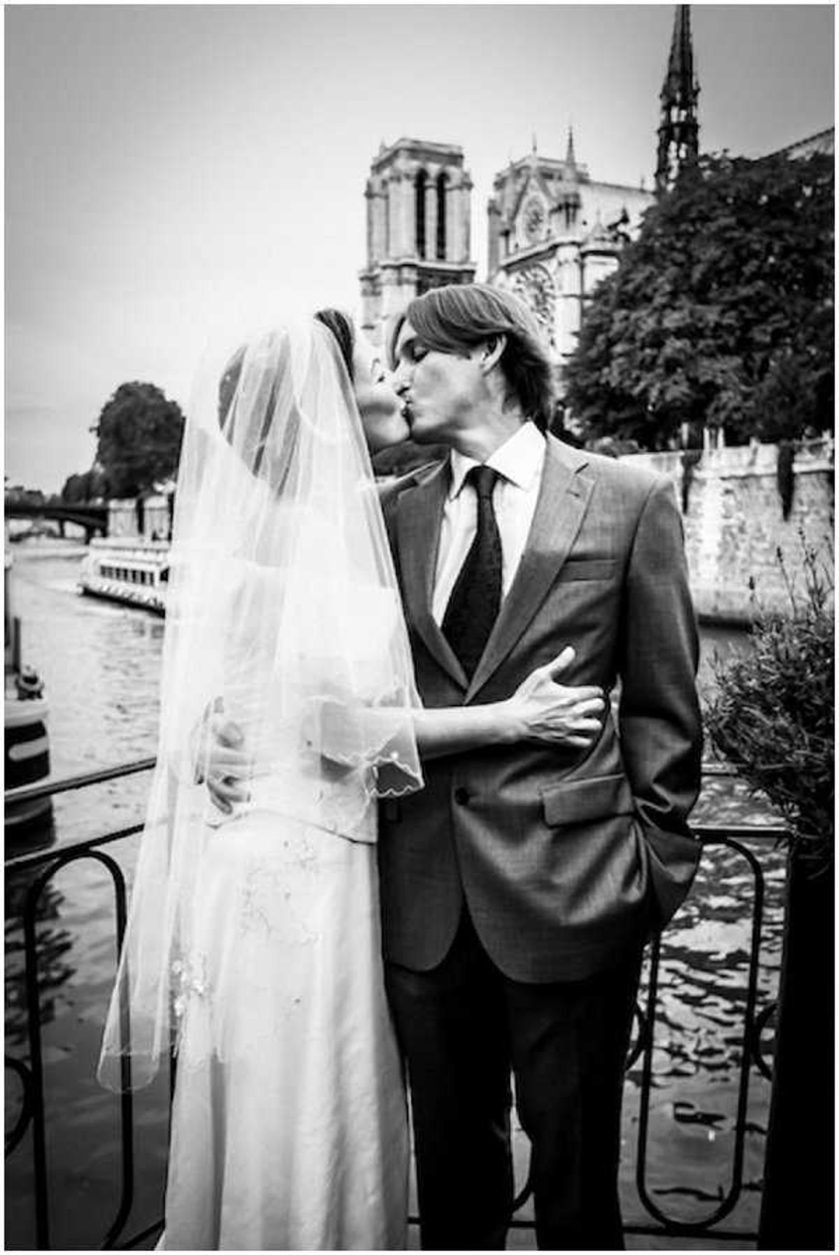 This black-and-white portrait shows a bride and groom kissing on the banks of the Seine River in Paris, with Notre-Dame Cathedral clearly visible in the background. The bride wears a full-length gown with embroidered detailing and a long cathedral-length veil, while the groom is dressed in a fitted suit jacket with a dark tie. The image has strong mid-tone contrast with bright highlights on the veil and darker tones in the background foliage and ironwork railing. The composition is a medium portrait shot taken at street level, with a bateau mouche boat visible on the river behind them.