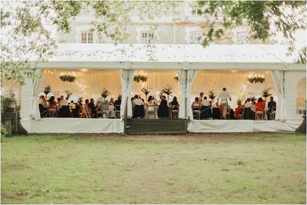 Wide exterior shot of a wedding reception taking place inside a large white marquee tent on the grounds of a French chateau, visible in the background. Approximately 40-50 guests are seated at tables inside the open-sided tent, which is decorated with warm pendant lighting, draped white fabric along the interior walls, hanging floral or greenery chandeliers, and tall tropical-style potted plants. Fairy lights are strung along the tent's exterior roofline. The decor palette is warm white and green with a classic garden party feel. The image is taken from across the lawn at dusk, giving a wide establishing shot that shows both the tent and the chateau facade behind it. Potential venue feature image.