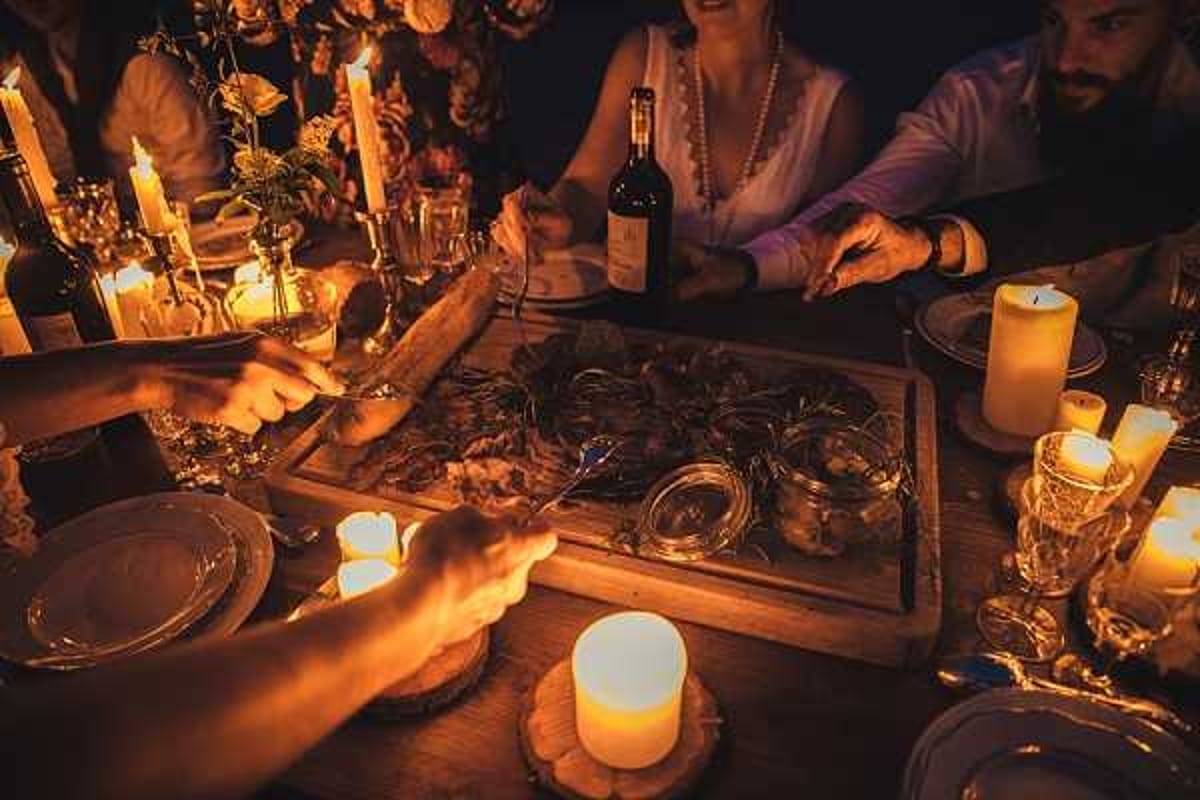 A wedding reception dinner scene shot from above at close range, showing guests gathered around a rustic wooden table sharing a communal charcuterie-style platter served on a large wooden board. The table is lit entirely by candlelight — pillar candles, taper candles in candleholders, and small votive tea lights — casting deep amber and warm golden tones across the scene. Visible table elements include crystal glassware, silver cutlery, ceramic plates, small glass jars with condiments, and a red wine bottle. Floral arrangements with dried and fresh blooms in muted tones are visible in the background. At least three guests are partially visible — a woman in white wearing a pearl necklace, a man in a light shirt, and another figure in dark clothing — all reaching toward the shared board. The overall styling is rustic and intimate, with the candlelight-only setting creating very dark shadows and high contrast across the image.