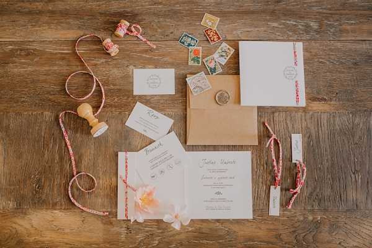 A flat lay of a wedding stationery suite arranged on a rustic wooden surface. The suite includes a main invitation card with script typography, an RSVP card, a small enclosure card, a kraft paper envelope sealed with a round wax seal, and a plain white outer envelope. Styling accents include a wooden rubber stamp, a spool of red and white patterned ribbon loosely arranged across the layout, several vintage decorative postage stamps in jewel tones, a small pink flower blossom, and two thin place card strips. The overall palette is white, kraft brown, and red with botanical accents, giving the suite a romantic vintage-postal aesthetic.