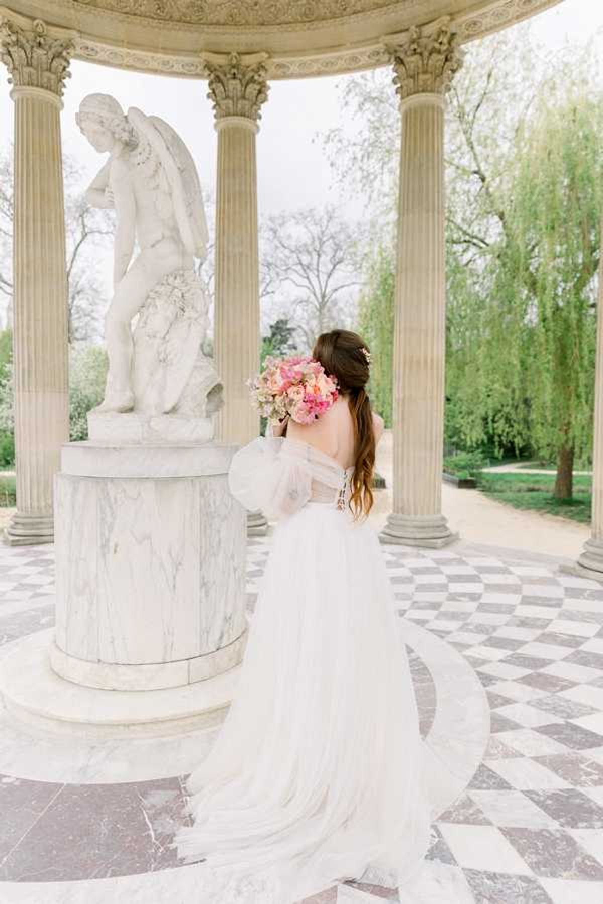 A bridal portrait taken from behind inside a neoclassical rotunda or pavilion, featuring tall Corinthian columns and a black-and-white diamond-patterned marble floor. The bride stands facing a large white marble angel sculpture on a veined marble pedestal, holding a lush bouquet of pink, coral, and blush blooms including what appear to be peonies and sweet peas. She wears an off-the-shoulder white tulle ballgown with sheer long sleeves and a full cathedral-length train spread across the floor, with floral hair accessories visible in her loose dark hair. The shot is a full-length portrait from behind, with a formal garden path and weeping willow trees visible through the columns in the background.