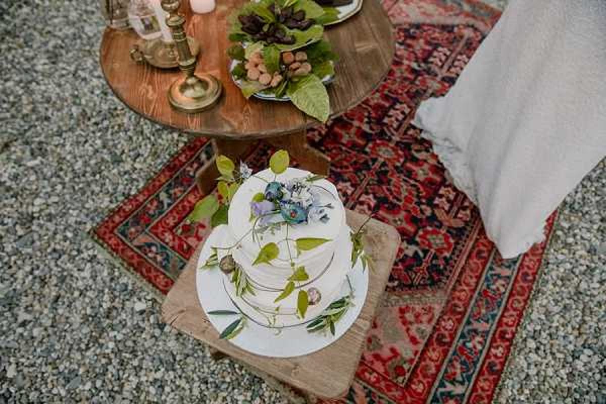Close-up detail shot of a two-tier semi-naked wedding cake placed on a rustic wooden stool, decorated with green foliage sprigs and small blue and purple flowers. The stool sits on a vintage red and navy patterned rug laid over a gravel surface, contributing to a boho outdoor styling aesthetic. In the background, a round wooden side table holds brass candlesticks and a decorative plate with berries and dark leaves. The edge of a white fabric garment is visible at the right of the frame.