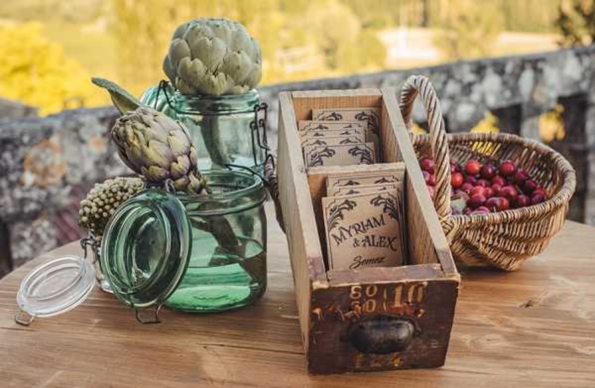 Close-up detail shot of a rustic wedding stationery and decor vignette displayed on a wooden table, likely at an outdoor cocktail hour or reception. A weathered wooden crate holds kraft paper seed packet wedding favors printed with 'Myriam & Alex Semée' (French for 'sown'), alongside teal-green glass mason jars, fresh artichokes used as decorative elements, and a woven wicker basket filled with red cherries or cranberries. The overall styling is rustic and nature-inspired, with an earthy, botanical theme carried through the seed packet favors, raw wood, and garden vegetables as decor accents.