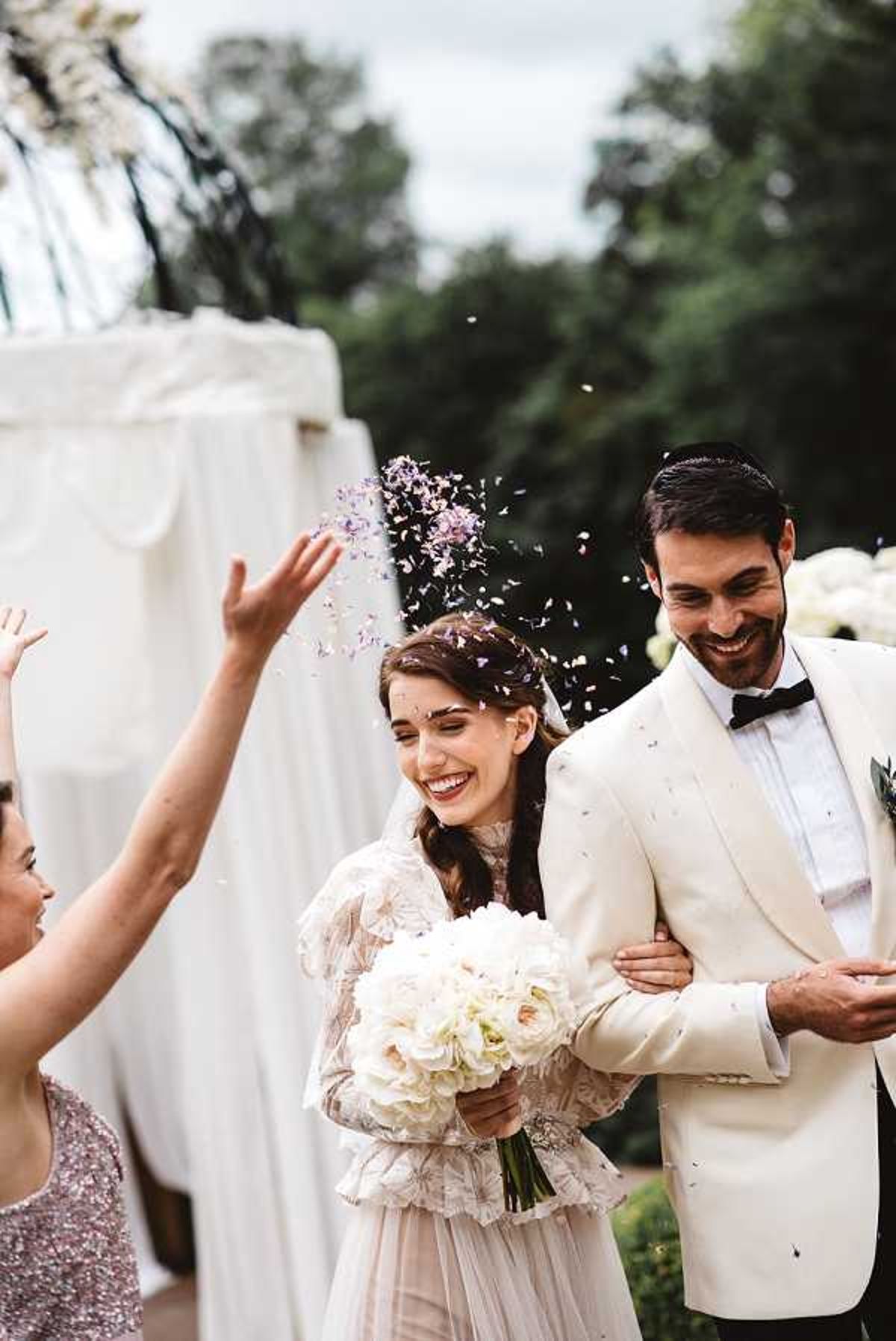 The bride and groom walk together as a guest throws lavender and white flower petal confetti over them during an outdoor ceremony exit. The bride wears a long-sleeved, embroidered blush-toned gown with a high neckline and carries a bouquet of white garden roses and hydrangeas, while the groom wears a cream dinner jacket with a black bow tie. Both are laughing, and a female guest in a rose-gold sequined dress is visible to the left mid-throw. The backdrop includes a white draped ceremony arch and white floral arrangements. The shot is a close-up portrait with shallow depth of field, captured in a natural, candid style.