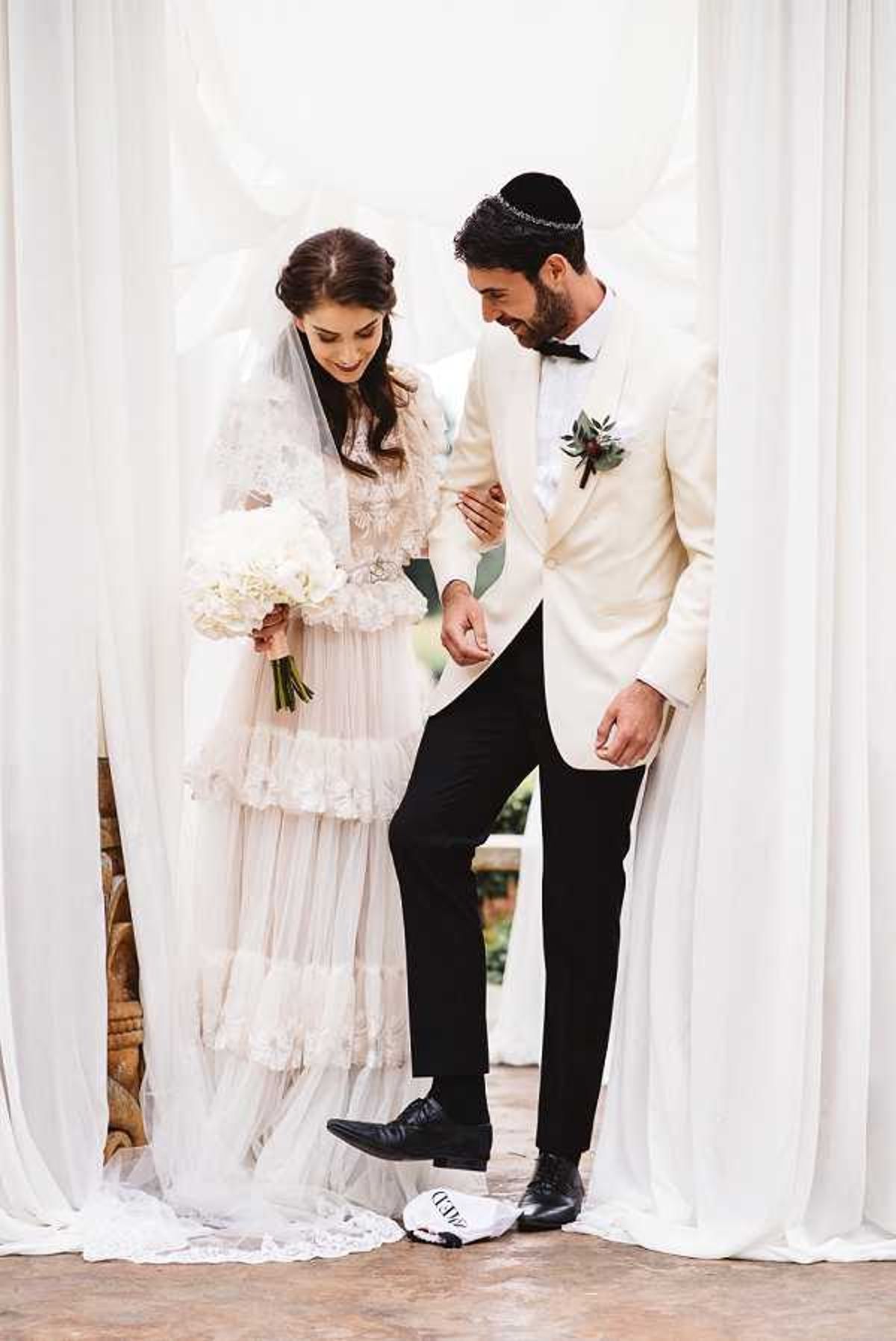 A couple is photographed at the moment of the Jewish glass-breaking tradition, with the groom about to stamp on a wrapped glass placed on the ground beneath a white fabric chuppah or draped curtain backdrop. The bride wears a tiered, layered ivory tulle and lace long-sleeve dress with a cathedral-length veil and holds a bouquet of white hydrangeas and roses. The groom wears a cream dinner jacket with black trousers, a black bow tie, a kippah with a decorative band, and a boutonniere featuring greenery and a dark flower. The shot is a full-length portrait with soft, even lighting, and the white draping frames the couple symmetrically.