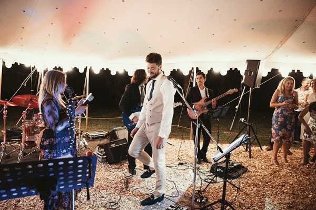 A live band performs during a wedding reception inside a large white sailcloth or marquee tent. The groom, wearing a cream three-piece suit with a dark tie and navy loafers, dances energetically at center stage in front of a microphone stand. The band includes a female vocalist in a floral dress, a guitarist in a dark jacket, and a drummer partially visible at the rear left, with amplifiers and music stands on a wood chip-covered ground floor. Several wedding guests in floral and formal attire watch and socialize along the right edge of the frame, with warm tent lighting and string lights visible along the tent's inner border. Wide shot capturing the full performance setup and guest interaction.