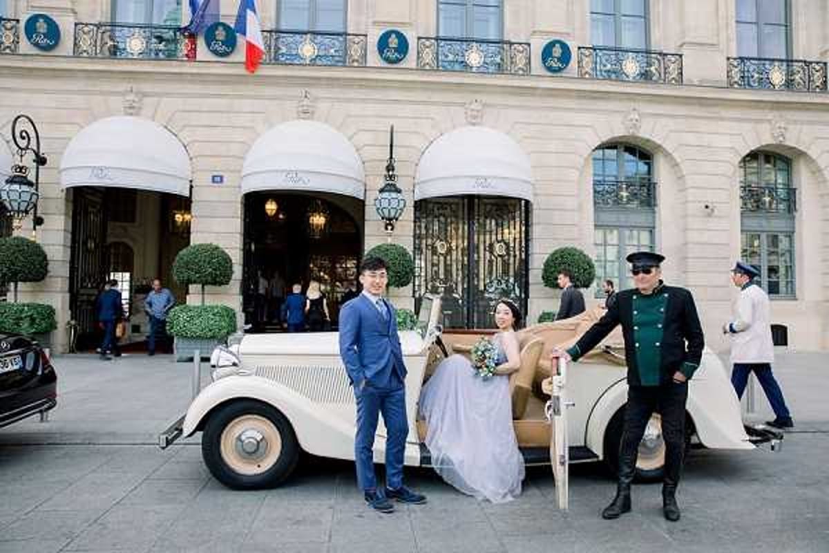 A couple poses with a vintage cream-and-tan open-top classic car in front of what appears to be the Ritz Paris on Place Vendôme, identifiable by the iconic striped dome awnings and the Ritz signage. The groom wears a navy blue suit and the bride is seated on the car in a layered tulle ball gown in a soft grey-blue tone, holding a small bouquet with blue and white flowers. A uniformed doorman in a dark green coat and peaked cap stands beside the car, while hotel staff in white uniforms and other bystanders are visible in the background. This is a wide portrait-style shot with the ornate Haussmann-style hotel façade, French flags, and manicured topiary as the backdrop. Potential venue feature image.