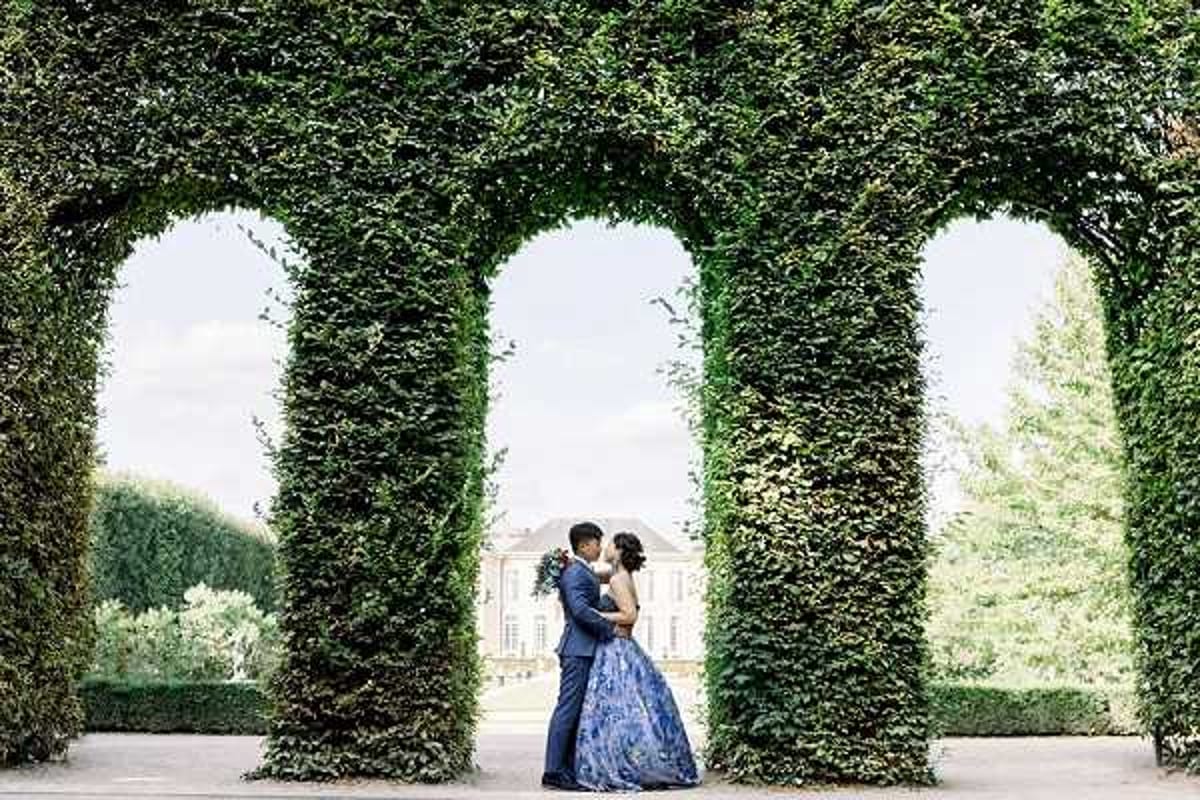 A couple poses for a portrait in the formal gardens of a French chateau, standing between tall ivy-covered hedge arches that frame the shot. The bride wears a floor-length blue and white floral ballgown, and the groom is dressed in a navy suit; they face each other in an embrace with a white bouquet visible. A classical chateau building is visible in the background through the central arch, indicating a historic estate venue. The image is a wide shot with the architectural hedge columns creating strong vertical symmetry around the couple. Potential venue feature image.